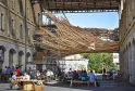 Outdoor courtyard in a historic stone building complex with visitors seated at wooden tables under a woven wooden canopy structure, featuring arches and modern architectural elements.