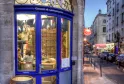 Cousin & Compagnie, a traditional wine shop with blue storefront and ornate golden awning on a Bordeaux street corner, displaying bottles in the window with a wooden barrel outside.