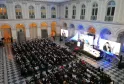 Large conference or gala event in the neoclassical Palais de la Bourse in Bordeaux, with hundreds of attendees seated facing a stage with multiple screens displaying speakers and presentations, ornate architecture with arched windows and columns visible i