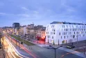 Modern white corner building of Seeko'o Hotel in Bordeaux at dusk, with historic architecture alongside, illuminated streets with light trails from traffic, and tram tracks visible
